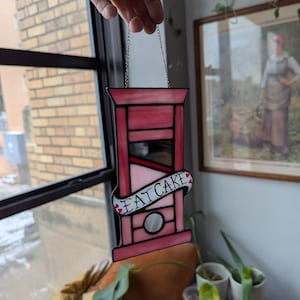 May include: A pink stained glass guillotine with a banner that reads "EAT CAKE" and two pink hearts. The guillotine is hanging from a silver chain.