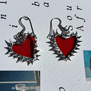 May include: A pair of heart-shaped earrings with a red center and a silver, spiky border. The earrings have a hook closure. The background includes text and a photograph.