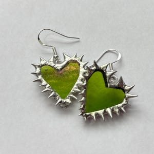 May include: A pair of heart-shaped earrings. Each earring features a bright green heart-shaped center framed by a silver, spiky border. The earrings have silver fish hook ear wires. The background is a plain white surface.