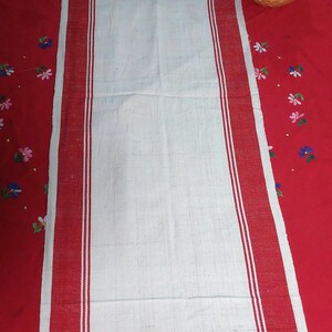 May include: A white rectangular table runner with red borders and two thin white stripes. The runner is placed on a red tablecloth with embroidered floral patterns. A yellow and blue ceramic pot with a plant is in the background.