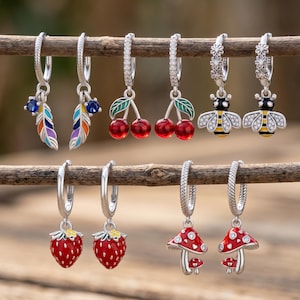 Può includere: Una collezione di orecchini a cerchio in argento con vari ciondoli. I ciondoli includono piume colorate, ciliegie rosse con foglie verdi, bombi, fragole rosse e funghi rossi con pois bianchi. Gli orecchini sono esposti su un ramo di legno.