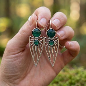 Può includere: Orecchini color argento con un design a raggiera. Ogni orecchino presenta una grande gemma verde scuro rotonda nella parte superiore e una più piccola al centro della raggiera. Gli orecchini sono tenuti da una mano.