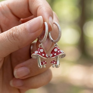 May include: Silver hoop earrings with red mushroom charms. The mushroom caps are red with white spots and small clear gems. The earrings are held up by a hand against a blurred green background.