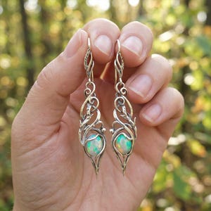 Puede incluir: Un par de pendientes colgantes plateados. Cada pendiente presenta un diseño ornamental y arremolinado con forma de lágrima en la parte inferior. Los pendientes tienen una piedra de ópalo ovalada con toques de verde y azul.