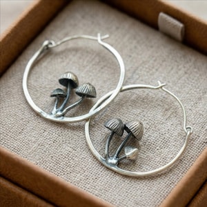 May include: Silver hoop earrings featuring a cluster of three detailed mushrooms. The earrings have a polished finish and are displayed in a brown jewelry box. The mushroom caps have textured detailing.