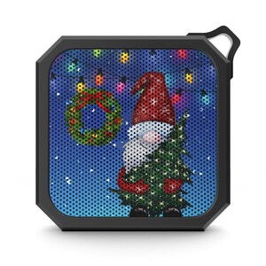 May include: A black, square portable speaker featuring a Christmas gnome design. The gnome wears a red hat and holds a Christmas tree. A wreath and string lights are also visible against a blue background.