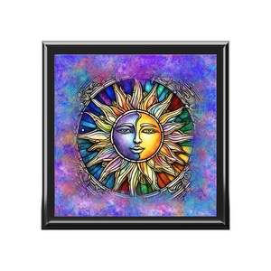May include: A square, framed artwork featuring a stylized sun with a face, split into yellow and purple halves. The sun is surrounded by colorful rays and a decorative border, set against a blue and purple background.