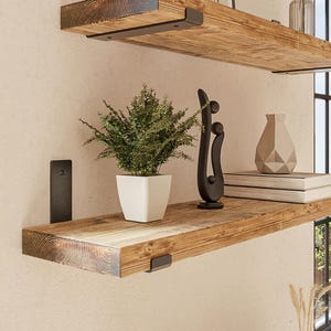 Può includere: Due mensole rettangolari in legno con venature naturali, montate a parete con staffe in metallo nero. Le mensole ospitano vari oggetti decorativi tra cui una pianta verde, libri, un vaso e delle sculture astratte.