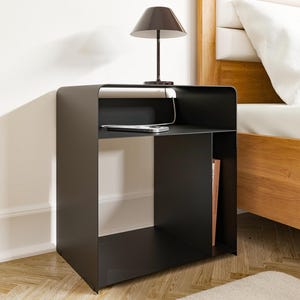 Könnte beinhalten: Moderner schwarzer Nachttisch mit integriertem Regal und kleiner Lampe. Ein Smartphone liegt auf dem Regal, verbunden mit einem Ladekabel. Der Tisch steht neben einem hölzernen Bettrahmen. Abmessungen: ca. 50 cm x 40 cm x 40 cm.