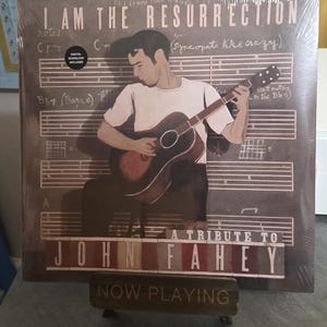 May include: A vinyl record titled "I Am The Resurrection: A Tribute to John Fahey." The album cover features an illustration of a man playing an acoustic guitar against a backdrop of musical notes. The words "Now Playing" are on a wooden stand.