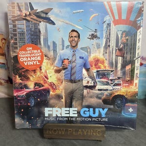May include: A vinyl record of the movie "Free Guy" featuring Ryan Reynolds. The album cover shows Reynolds holding a coffee cup in a city scene with explosions and aircraft. The text "ON COLLECTIBLE TRANSLUCENT ORANGE VINYL" is visible.