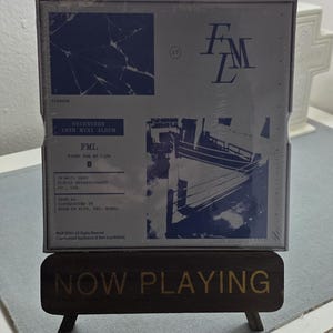 May include: A sealed album titled "FML" by Seventeen, featuring a blue and white design with a ship illustration. The album is displayed on a wooden stand that reads "NOW PLAYING" in gold lettering.