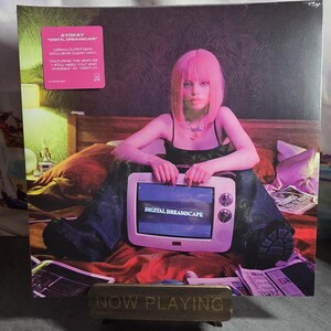 May include: A vinyl record titled "Digital Dreamscape" by Ayokay, with a pink sticker. The album cover features a person with pink hair holding a vintage television. The TV screen displays "Digital Dreamscape". A "Now Playing" sign is in the foreground.