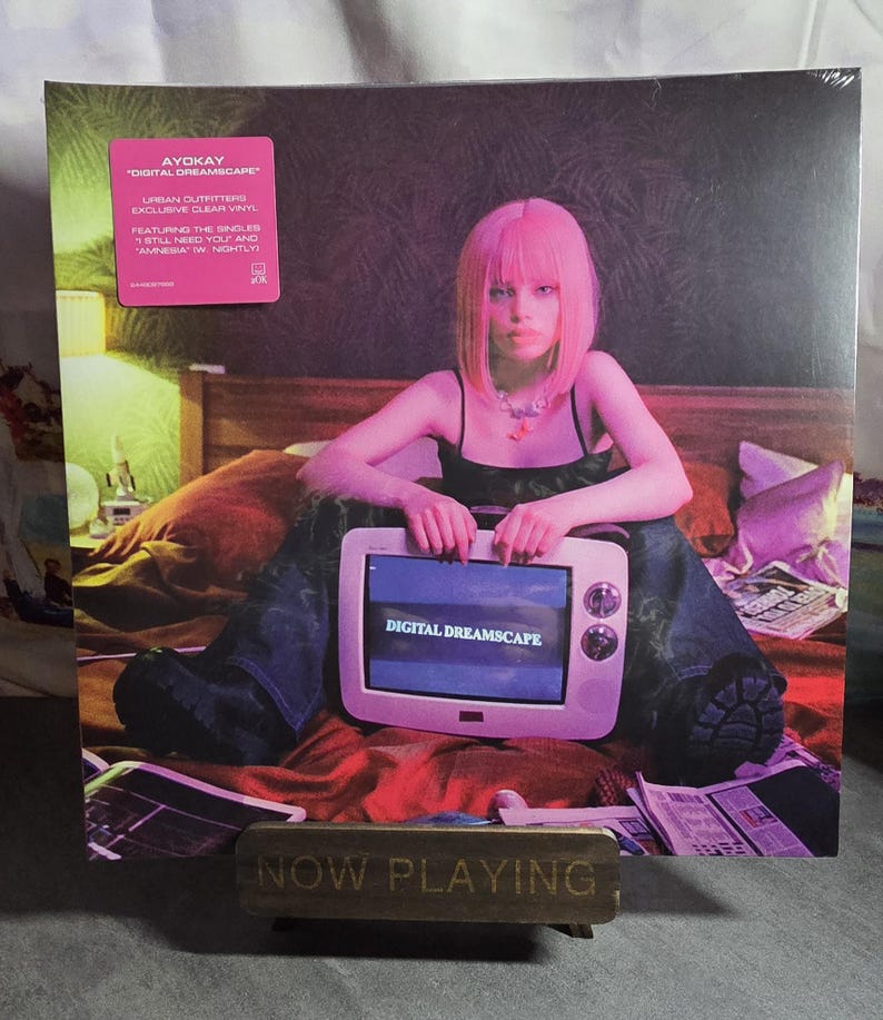 May include: A vinyl record titled "Digital Dreamscape" by Ayokay, with a pink sticker. The album cover features a person with pink hair holding a vintage television. The TV screen displays "Digital Dreamscape". A "Now Playing" sign is in the foreground.