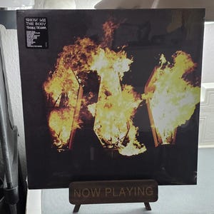 May include: A vinyl record album with a dark cover featuring three coffin shapes engulfed in flames. The album is titled "Show Me The Body" with the text "Trouble The Water" visible. A wooden stand below reads "NOW PLAYING".