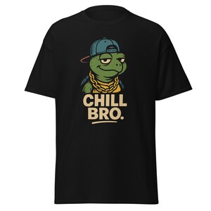 Könnte beinhalten: Schwarzes T-Shirt mit einer Zeichentrickschildkröte, die eine blaue Kappe und eine goldene Kette trägt. Die Schildkröte hat einen entspannten Ausdruck. Der Text "CHILL BRO." ist in einer stilisierten Schriftart unter der Schildkröte gedruckt.