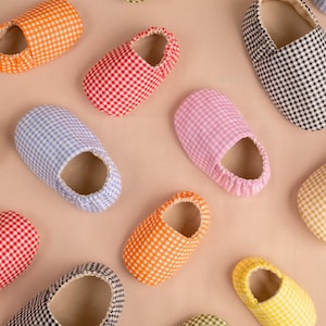 May include: A collection of colorful gingham slippers in various shades, including red, blue, pink, green, yellow, orange, and black. The slippers are arranged on a light pink surface, showcasing their soft, fabric construction and open-toe design.