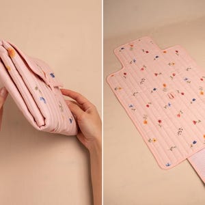 Puede incluir: Un cambiador acolchado rosa con un estampado floral. El cambiador est&aacute; doblado en la primera imagen y desplegado en la segunda, revelando una extensi&oacute;n forrada con cuadros vichy. Est&aacute; decorado con flores bordadas en varios colores.