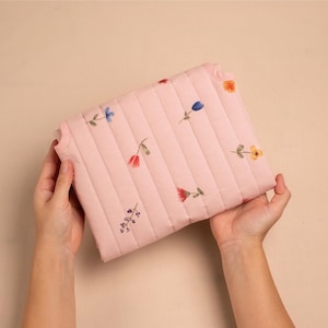 Puede incluir: Un bolso de mano acolchado rosa con un estampado floral. El bolso rectangular tiene costuras verticales y flores bordadas en rojo, azul y amarillo. El fondo es de un color neutro.