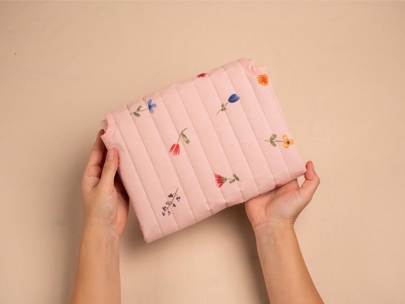 Puede incluir: Un bolso de mano acolchado rosa con un estampado floral. El bolso rectangular tiene costuras verticales y flores bordadas en rojo, azul y amarillo. El fondo es de un color neutro.