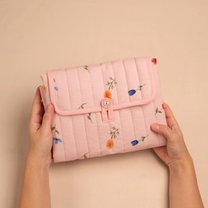 Puede incluir: Un bolso de mano acolchado rosa claro con cierre de solapa y bot&oacute;n. El bolso de mano est&aacute; decorado con peque&ntilde;os estampados florales en azul, naranja y verde. El bolso de mano est&aacute; sostenido por dos manos.