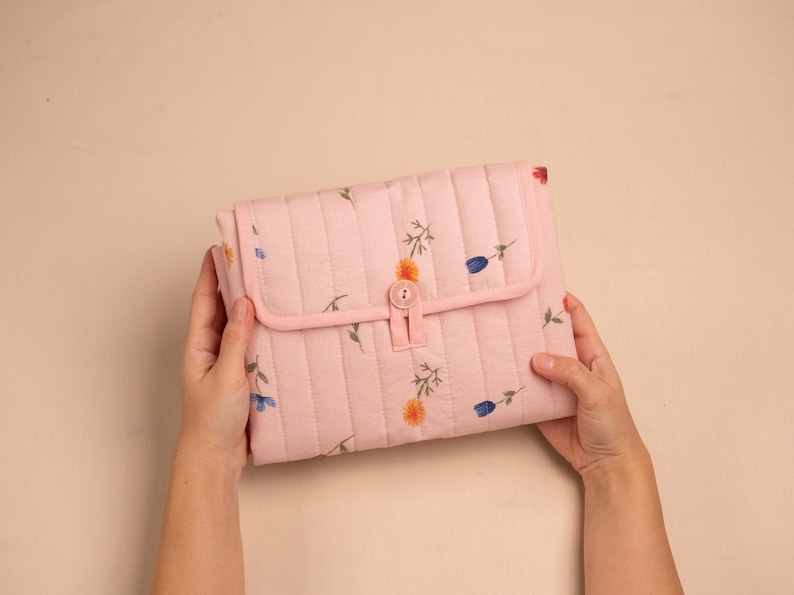 Puede incluir: Un bolso de mano acolchado rosa claro con cierre de solapa y bot&oacute;n. El bolso de mano est&aacute; decorado con peque&ntilde;os estampados florales en azul, naranja y verde. El bolso de mano est&aacute; sostenido por dos manos.