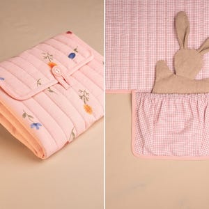 Puede incluir: Un cambiador acolchado rosa con un estampado floral y cierre de bot&oacute;n. La imagen tambi&eacute;n muestra una funda de cambiador de cuadros rosas y blancos con un bolsillo y un conejito de peluche.
