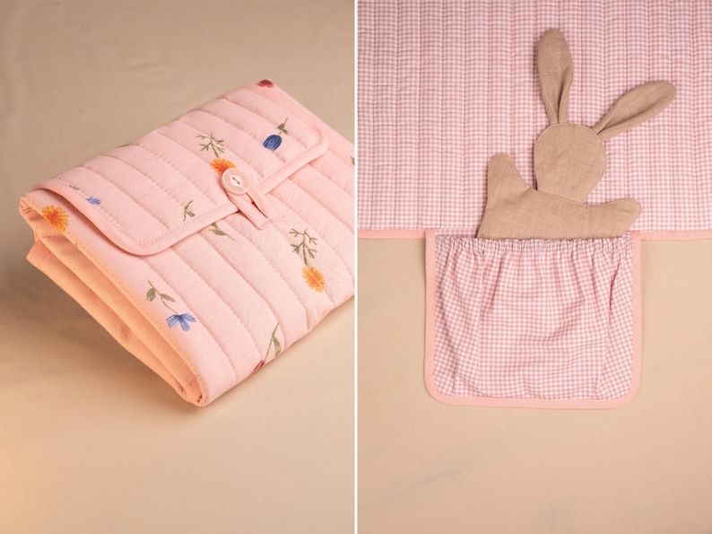 Puede incluir: Un cambiador acolchado rosa con un estampado floral y cierre de bot&oacute;n. La imagen tambi&eacute;n muestra una funda de cambiador de cuadros rosas y blancos con un bolsillo y un conejito de peluche.