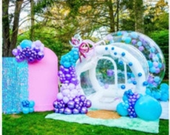 May include: A large clear inflatable bubble with purple, blue and white balloons surrounding it. The bubble is decorated with a white bow and is set up outdoors on a green lawn.