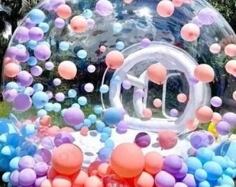 May include: A transparent inflatable bubble tent filled with pastel-coloured balloons in shades of pink, purple, and blue. The tent is situated outdoors on grass, creating a whimsical and festive atmosphere for a celebration.