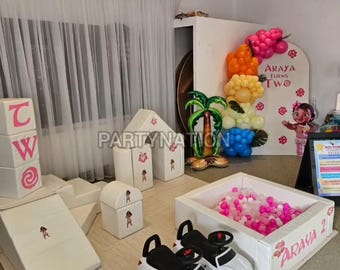 May include: A children's party setup with white and pink decorations, including a ball pit, foam blocks, and toy cars. The setup includes a balloon arch and a sign that says "ARAYA TURNS TWO". The party is themed around a cartoon character.