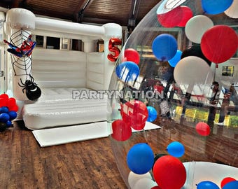 May include: A white inflatable bouncy castle with a Spiderman theme and a large clear inflatable bubble filled with red, blue and white balloons. The bouncy castle has a black spider web and a black spider on the front. The bubble is sitting on a wooden floor. The text "Partynation42@gmail.com", "Insta: Partynation42", and "# 07944988299" is visible in the image.
