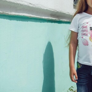 Op de afbeelding: Een jong meisje draagt een wit T-shirt met een roze en gele cartoonkuiken met een zonnebril en een roze strik. Het shirt zegt "One Cute Chick". Het meisje draagt een blauwe jeans en een zonnebril.