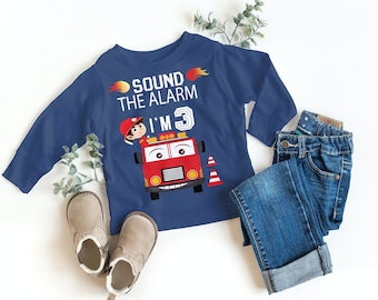 May include: A blue long-sleeve shirt with a red fire truck graphic and the text "Sound the Alarm I'm 3". The shirt is on a white surface with a pair of brown boots and blue jeans.