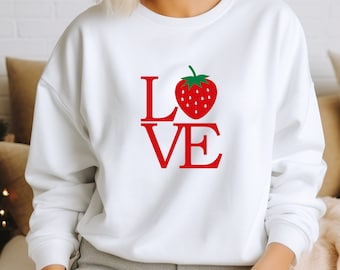 Könnte beinhalten: Weißer Pullover mit einem roten "LOVE"-Motiv, das eine rote Erdbeere mit grünen Blättern in der Mitte des "O" zeigt.