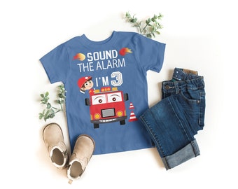 May include: A blue t-shirt with a red fire truck and a firefighter on it. The shirt says "Sound the Alarm I'm 3".