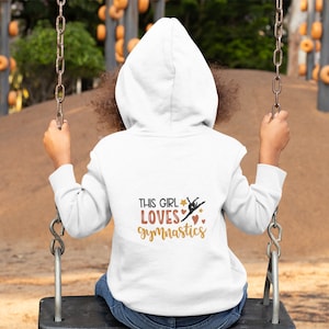 Könnte beinhalten: Weißer Kapuzenpullover mit dem Text "This girl loves gymnastics" und einer Silhouette einer Turnerin in einem Sternenstrahl.