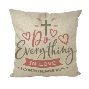 Könnte beinhalten: Ein beiger Kissenbezug mit einem braunen Kreuz und dem Text "Do Everything in Love" mit zwei rosa Herzen auf beiden Seiten des Wortes "Do". Unter dem Text befindet sich ein rotes Band mit dem Text "IN LOVE" und dem Bibelvers ". 1 Korinther 16:14."