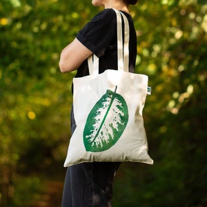Könnte beinhalten: Eine Person trägt eine weiße Einkaufstasche mit einem grünen Blattdesign. Die Tasche ist aus Canvas und hat zwei lange Henkel. Die Person steht in einer Waldumgebung.