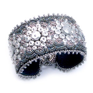 Peut inclure: Un large bracelet manchette argenté orné de perles. Le bracelet est richement orné de perles et de paillettes argentées de différentes tailles, créant une surface texturée. L'intérieur est doublé d'un tissu bleu foncé.