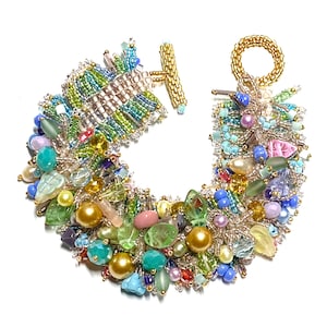 May include: A colourful beaded bracelet with a mix of green, blue, pink, and gold beads. The bracelet has a toggle clasp and a gold-toned ring. The design incorporates various shapes and sizes of beads, creating a unique accessory.