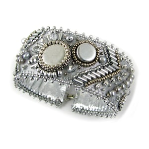 May include: A silver bracelet with a beaded design featuring two round silver accents. The bracelet is made of fabric and has a clasp closure.