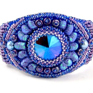 May include: A blue beaded bracelet with a large iridescent blue crystal in the center. The bracelet is decorated with smaller beads and pearls.