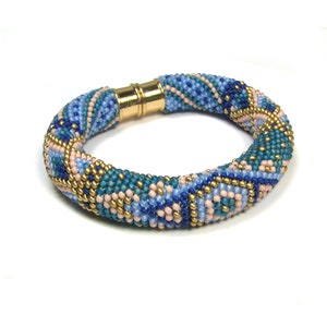 May include: A beaded bracelet featuring a geometric pattern in shades of blue, pink, and gold. The bracelet is crafted with small seed beads and has a magnetic clasp.