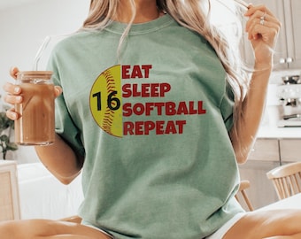 May include: A woman wearing a green t-shirt with the text "Eat Sleep Softball Repeat" and a softball graphic with the number 16. She is sitting on a white surface and holding a glass of iced coffee.