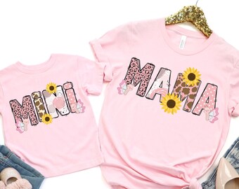 May include: Pink t-shirts with the words "Mama" and "Mini" in a colorful, patterned font. The shirts feature butterflies, sunflowers, and animal print designs.