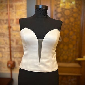 Puede incluir: Un top corsé de satén blanco con escote corazón y un panel transparente. La parte superior se muestra en un maniquí negro, mostrando su elegante diseño. El corsé es una pieza clásica de ropa formal.