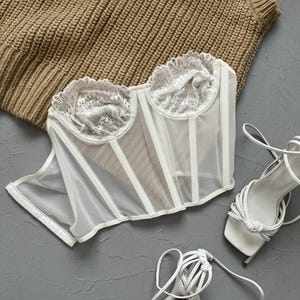 Puede incluir: Corsé blanco de encaje y malla con escote de corazón. El corsé está sobre un fondo gris con un suéter de punto marrón y tacones blancos de tiras.