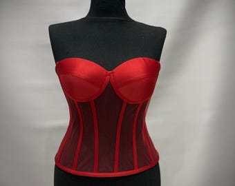 May include: A red satin corset with a sweetheart neckline and boning detail. The corset is displayed on a black mannequin. The bodice features a sheer mesh paneling and a structured silhouette, offering a classic and elegant look.