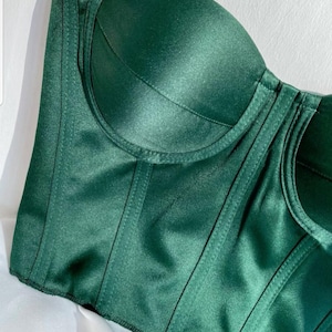 May include: A close-up of a dark green satin corset top. The bustier features structured boning and seams, creating a defined shape. The fabric has a subtle sheen, and the design is simple and elegant.